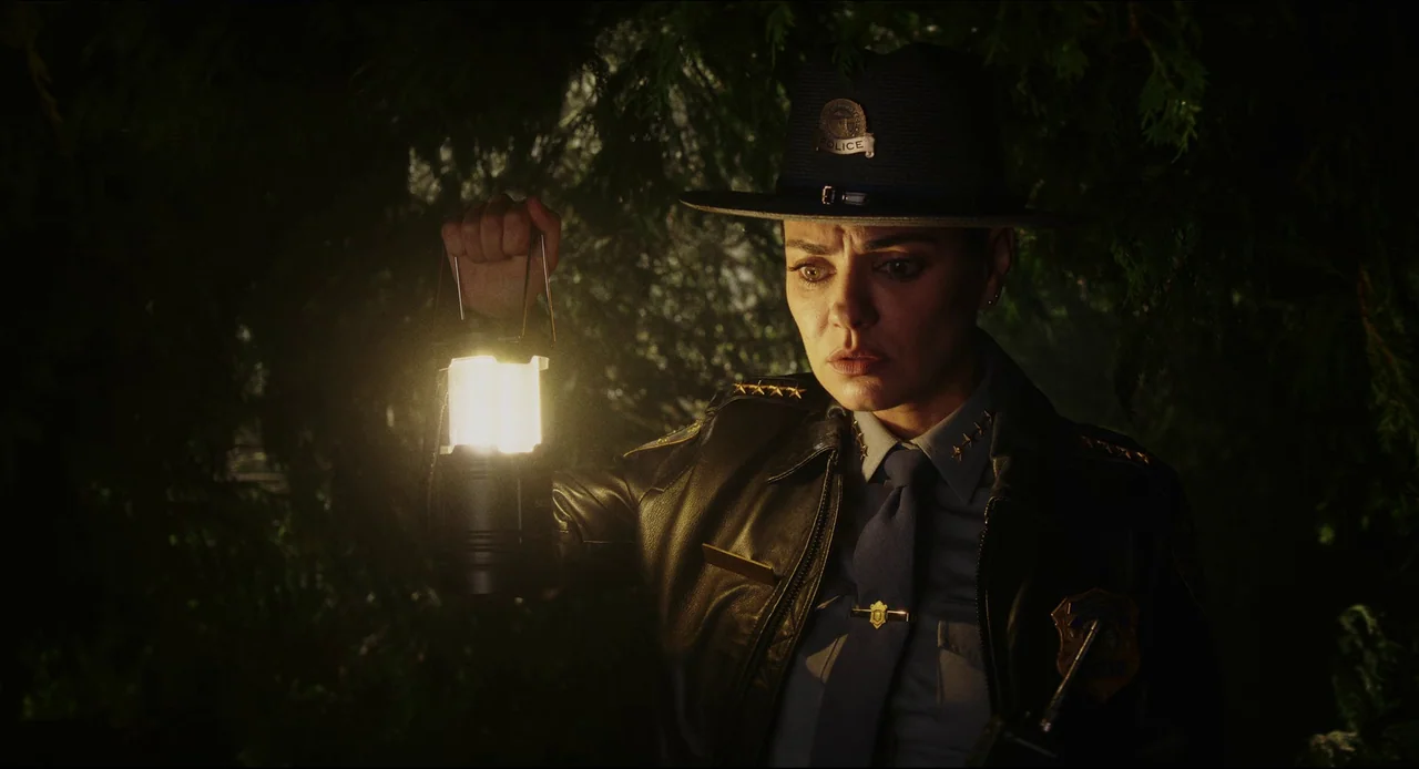 Wake Up Dead Man (2025) movie still: medium — A police officer stands surrounded by dark, dense coniferous trees in the night. She holds a portable…