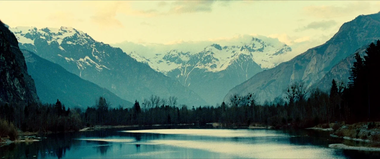 The Five Devils (2022) movie still: establishing — A wide mountain valley features a calm lake flanked by tall evergreen pine trees. Snow-capped…