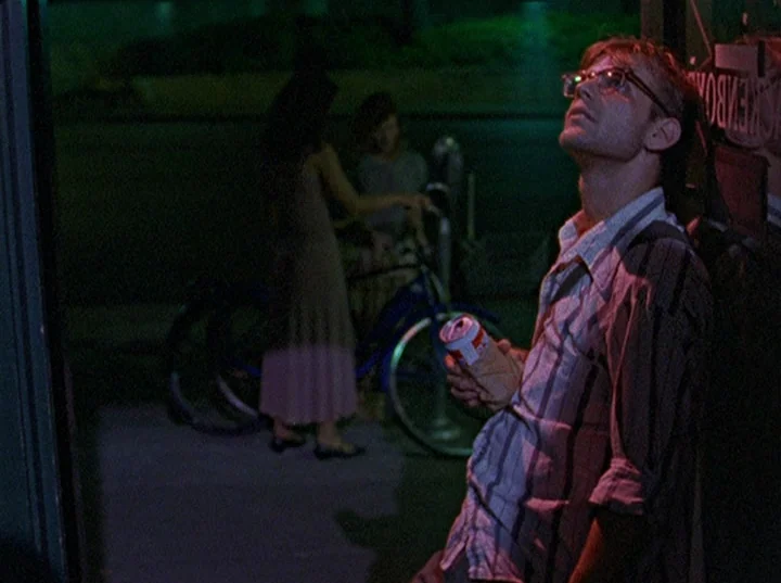 Slacker (1991) movie still: medium — A night exterior showing the entrance to a store illuminated by vibrant neon signage. A man stands in the…