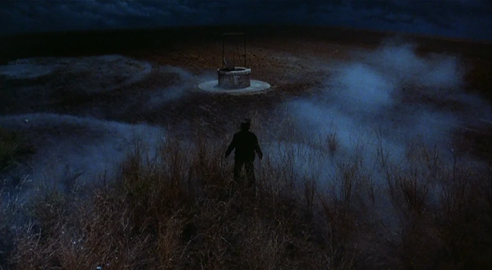 The Voice of the Moon (1990) movie still: wide — A dark night landscape features a stone well standing in an open grassy field. A lone figure stands…