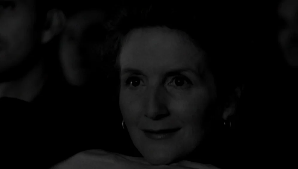 The Tango Lesson (1997) movie still: closeup — A dark, dimly lit theater space with blurred figures in the background. A woman sits in the…
