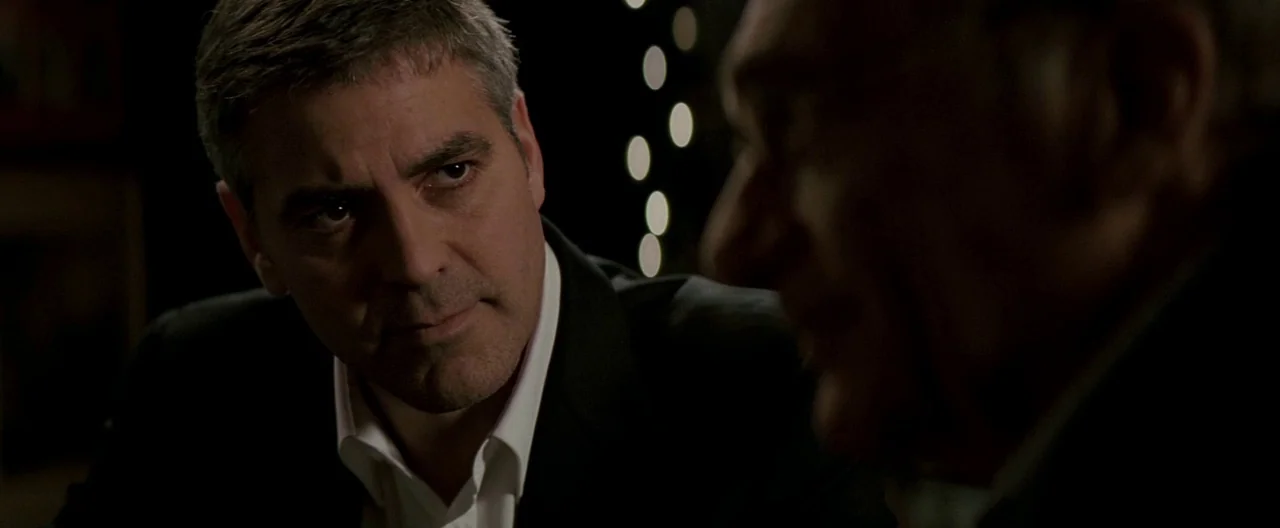 Michael Clayton (2007) movie still: closeup — Two men sit in a dark room with bokeh lights visible in the background. George Clooney faces his…