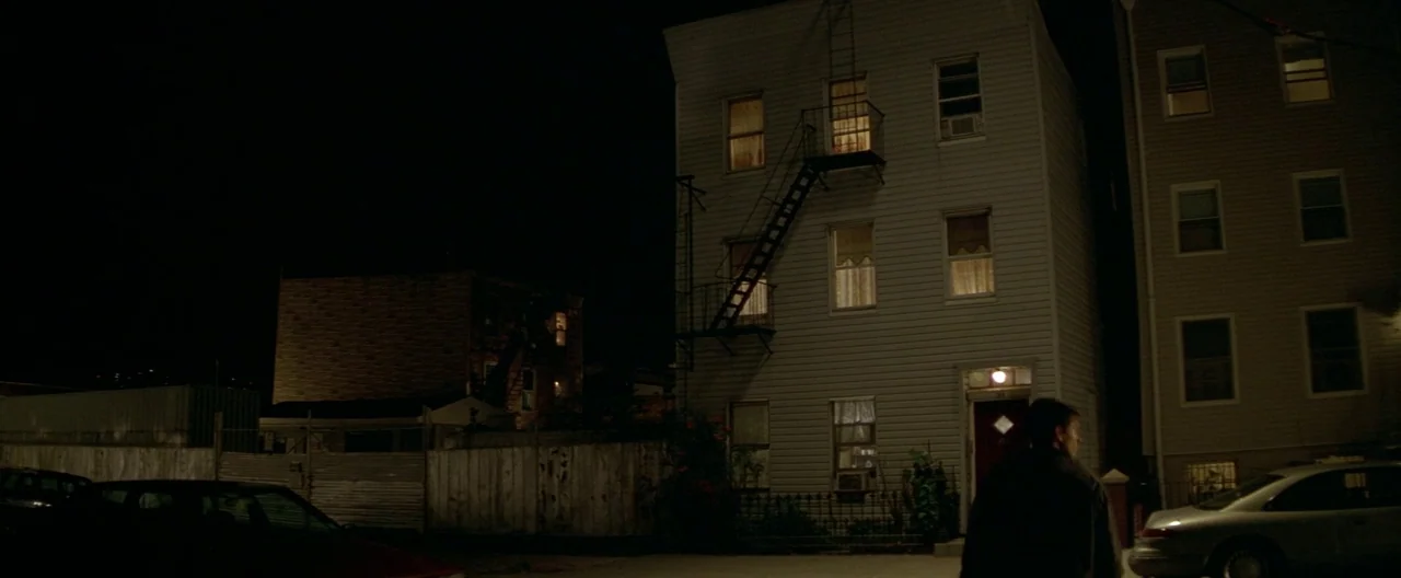 The Yards (2000) movie still: wide — A residential street at night features multi-story apartment buildings with yellow lit windows. A lone man in a…