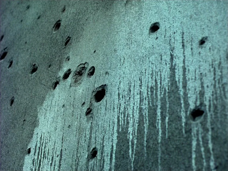 Notre Musique (2004) movie still: closeup — A weathered concrete wall is covered in multiple dark impact holes and mineral stains. The vertical…