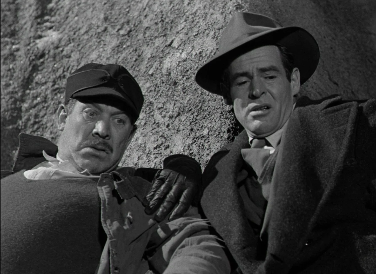 On Dangerous Ground (1951) movie still: medium — Two men huddle closely together against a rough, textured stone wall in bright daylight. One man…