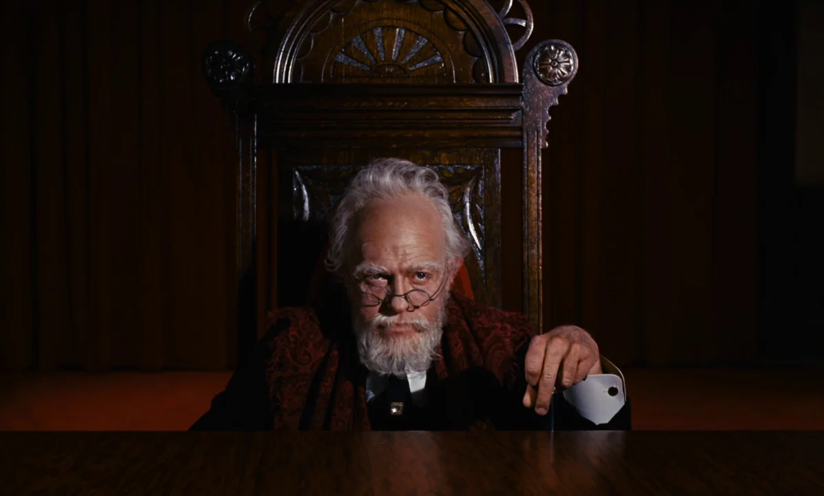 Mary Poppins (1964) movie still: medium — An elderly man sits at a desk in a room with dark red curtains. He faces forward from a high-backed wooden…