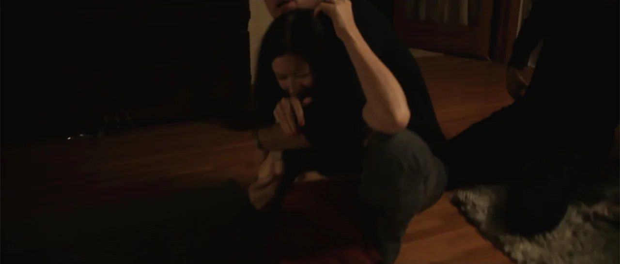La coherencia del caos movie still: medium — A dimly lit room features a hardwood floor and a partial view of furniture. A woman in a black shirt…