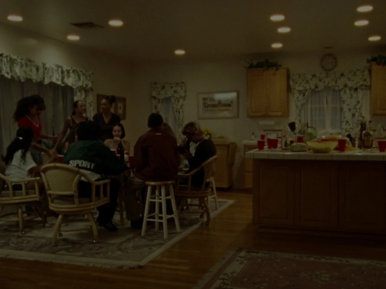 Mid90s (2018) movie still: wide — A residential kitchen features wooden cabinets and floral curtains over a window. Several teenagers sit around a…