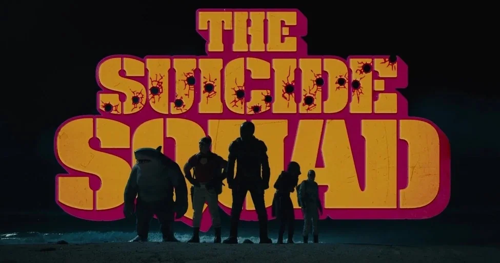The Suicide Squad (2021) movie still: wide — A group of five figures stand on a dark, wet shoreline at night. Behind them, enormous bold yellow and…