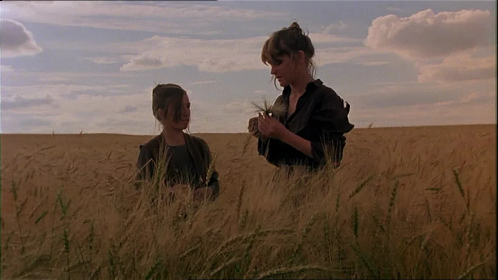 Days Of Heaven (1978) movie still: medium — A vast field of mature wheat stretches toward a pale sky with scattered clouds. Two women stand…