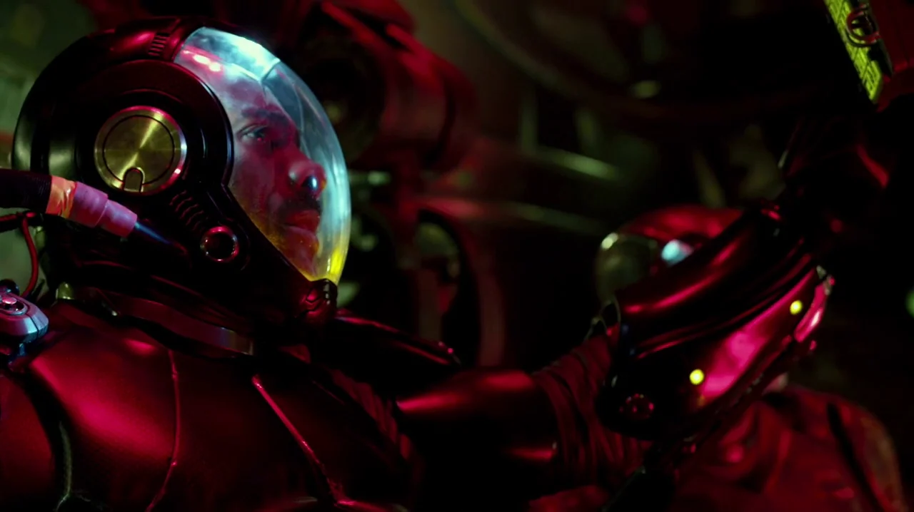 Pacific Rim (2013) movie still: closeup — A dark, cramped interior of a high-tech vessel illuminated by saturated red neon light. A person wearing a…