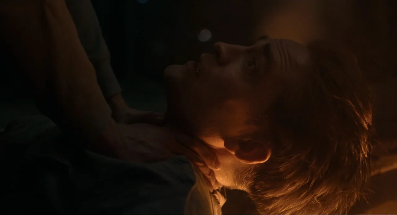 Mickey 17 (2025) movie still: closeup — A dark indoor space is lit by a warm, low-intensity light source. A person's hands grip the throat of a man…