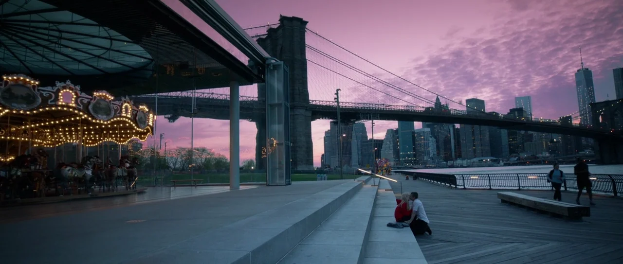 Nerve (2016) movie still: wide — A wide view of a modern waterfront park at dusk with the Brooklyn Bridge visible. A couple sits on concrete steps…