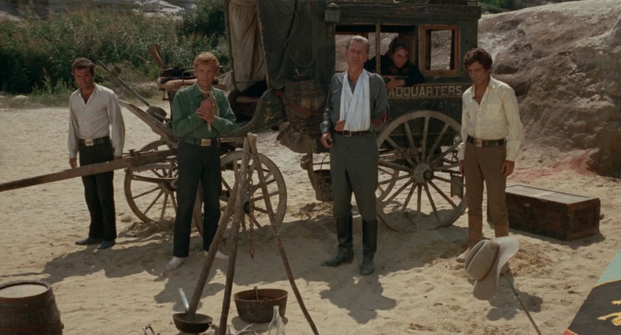 The Hellbenders (1967) movie still: fullbody — A weathered wooden wagon sits on a sandy, sun-baked landscape with sparse vegetation. Four men and…