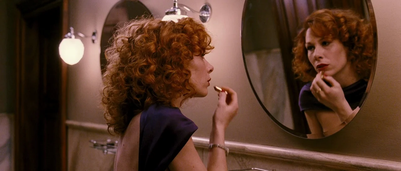 Black Book (2006) movie still: medium — A woman stands before a vanity with two circular mirrors mounted on a wall. She applies lipstick to her…