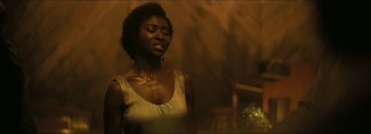 Sinners (2025) movie still: medium — A dimly lit room features wooden paneled walls and indistinct furniture in the background. A woman in a…
