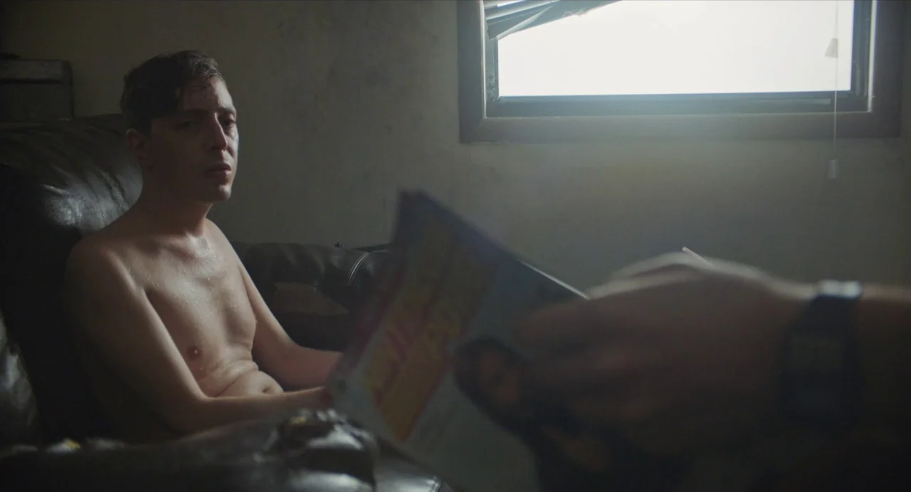 Relaxer (2018) movie still: medium — An interior space contains a dark leather armchair and a window with blinds. A shirtless man sits on the chair…