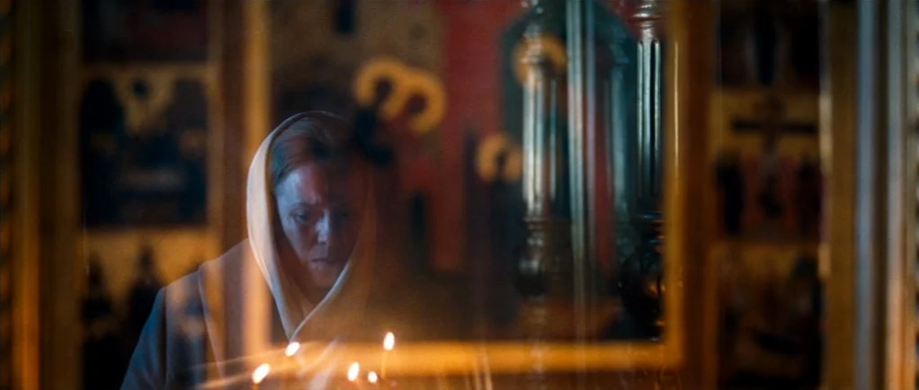 Elena (2011) movie still: medium — A dimly lit church interior with gold-framed icons visible behind glass. An older woman stands before the icons…