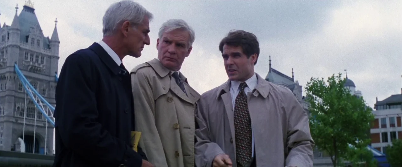 Mission: Impossible (1996) movie still: medium — Three men stand before Tower Bridge on an overcast day. They are engaged in an active conversation.