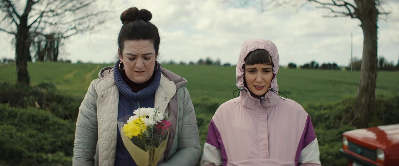 Extra Ordinary (2019) movie still: medium — An open rural field with sparse trees and an overcast sky background. Two women stand together in the…