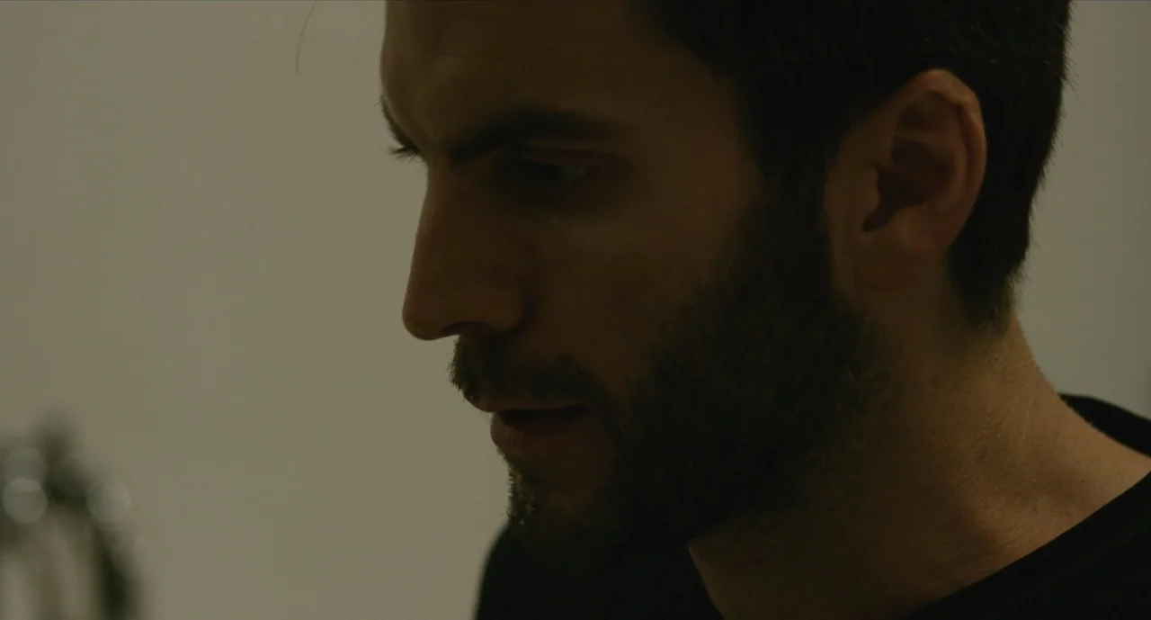 We Are Your Friends (2015) movie still: closeup — A plain, dimly lit interior space with a muted off-white wall background. A bearded man in a black…