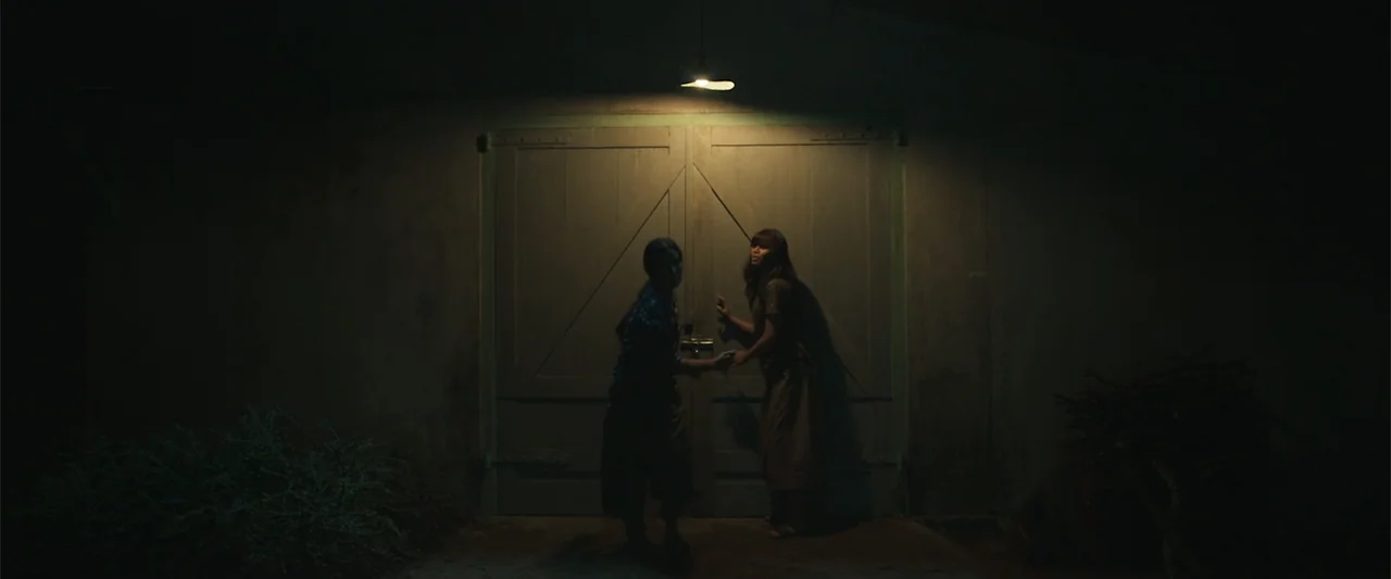 The Unholy (2022) movie still: fullbody — Large wooden double doors are illuminated by a single pendant light at night. Two women reach for the…
