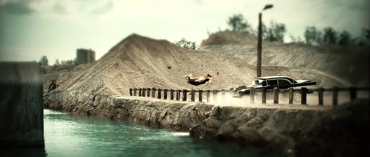 Daybreakers (2009) movie still: wide — A gravel quarry with large piles of excavated rock borders a turquoise water channel. A person in a suit is…
