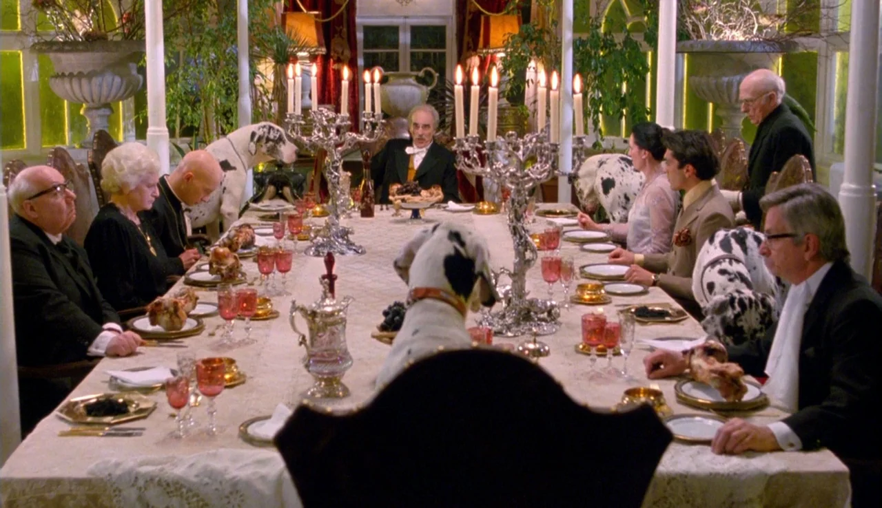 The Rainbow Thief (1990) movie still: wide — A long dining table is occupied by people and dalmatians in a formal space. A man sits at the head of…