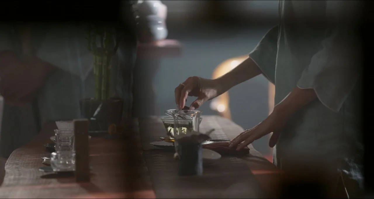 Five Blind Dates (2024) movie still: medium — A wooden table holds glass tea wares and a small green plant in a pot. A woman in light clothing uses…