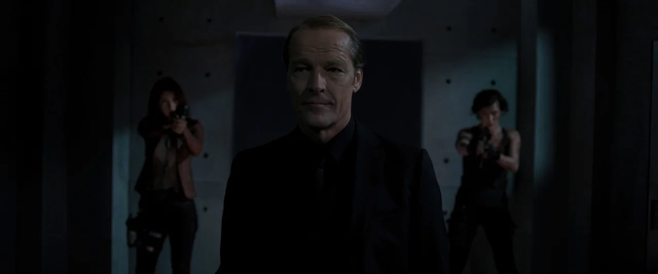 Resident Evil: The Final Chapter (2016) movie still: medium — A dark concrete corridor with visible bolt markings on the wall panels. A middle-aged…