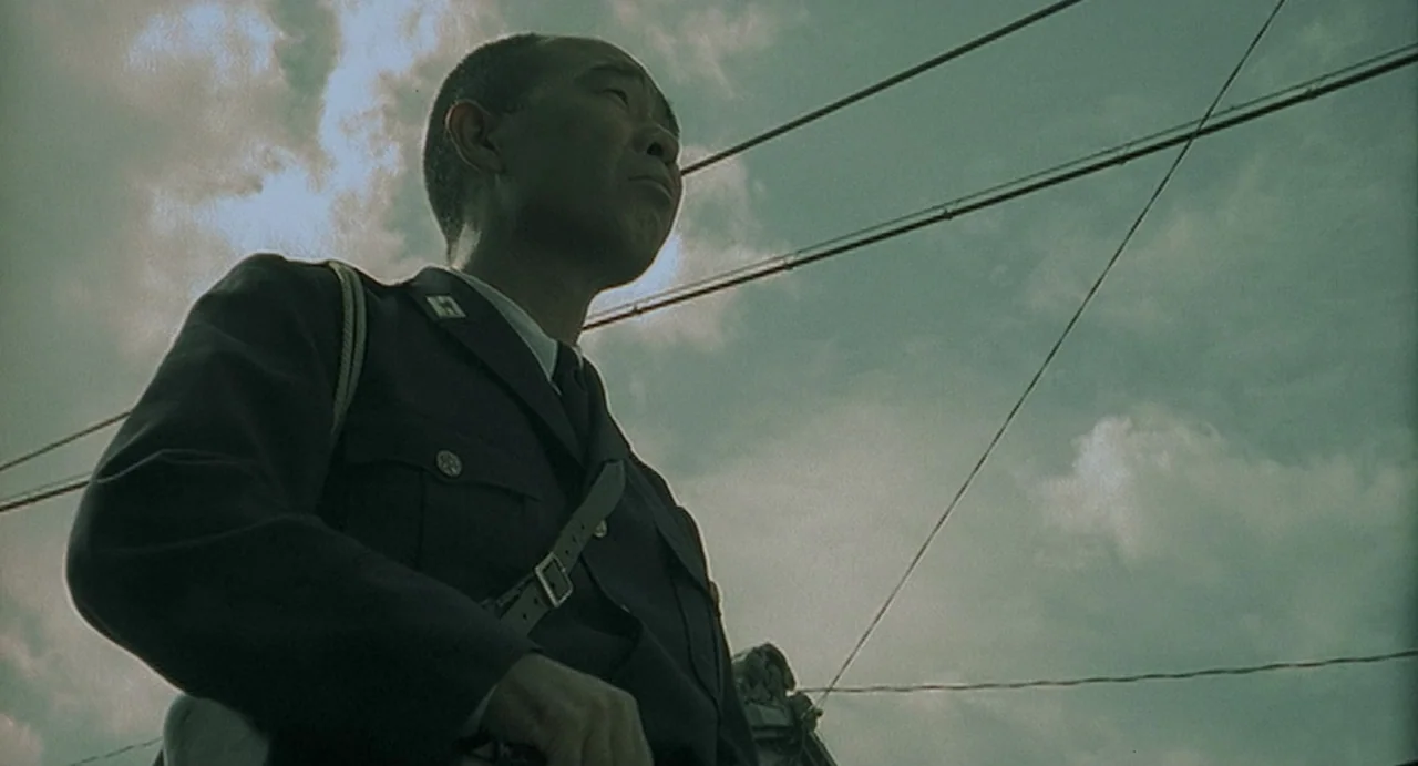 Uzumaki (2000) movie still: medium — An outdoor area features power lines visible against an overcast, cloudy sky. A man wearing a dark military…