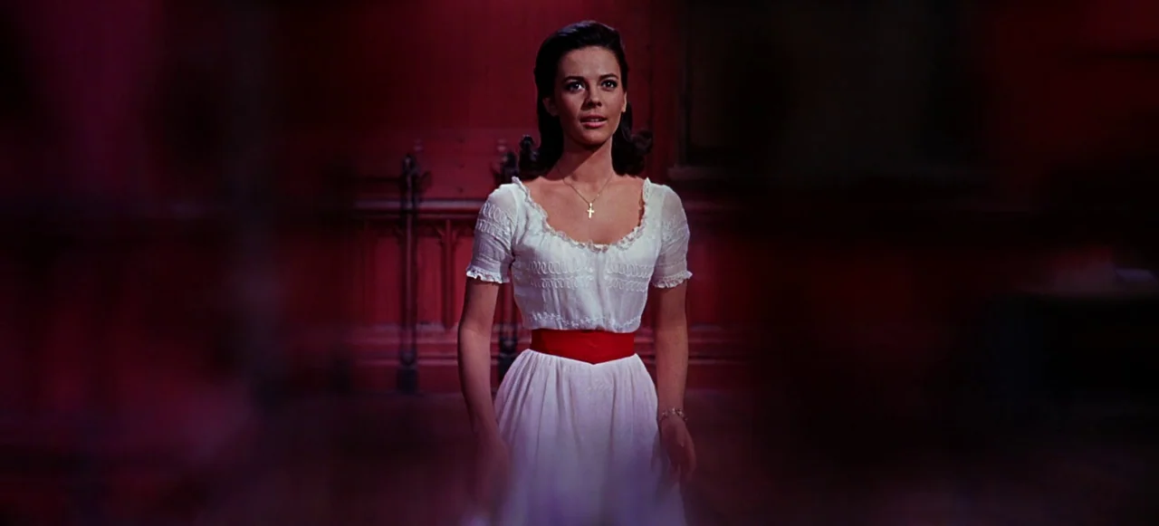 West Side Story (1961) movie still: medium — A high school gymnasium interior features dark red paneled walls and dim lighting. A woman stands alone…
