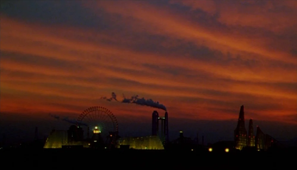Gozu (2003) movie still: establishing — A silhouette of an amusement park and industrial facilities stands against a vibrant sunset sky. Thin dark…