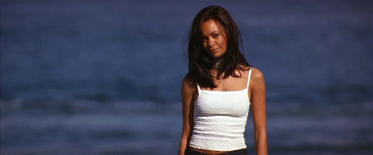 Mission: Impossible II (2000) movie still: medium — A woman stands before the blurred blue waves of the ocean at the beach. She faces forward while…