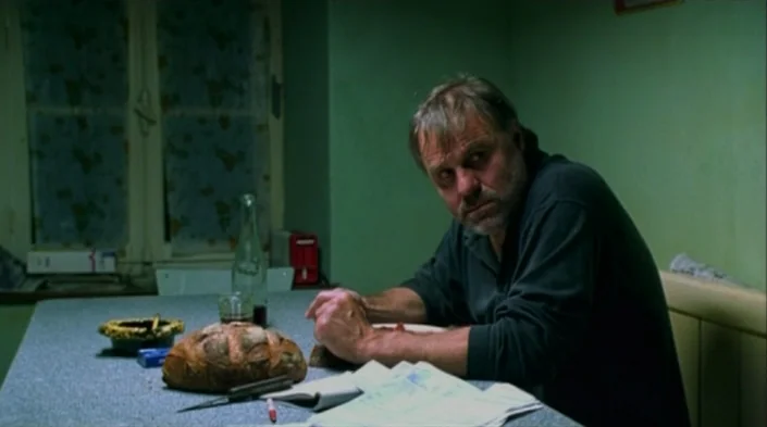 Code Unknown (2000) movie still: medium — A man sits at a table in a room with faded green walls. He is resting his hands on a surface covered with…