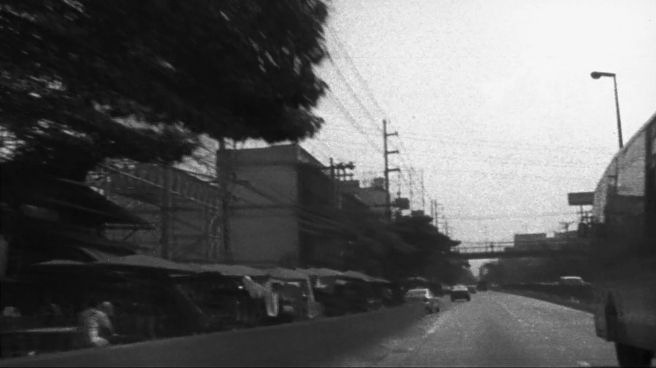 Mysterious Object at Noon (2000) movie still: wide — A roadside area features multiple small canopy stalls beside an open highway. Utility poles and…