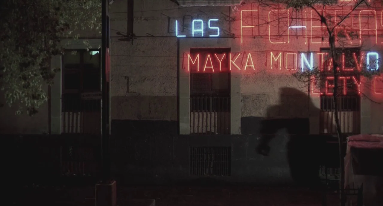 Santa Sangre (1989) movie still: wide — A nighttime street scene displays a building wall decorated with glowing red and blue neon. The neon text…