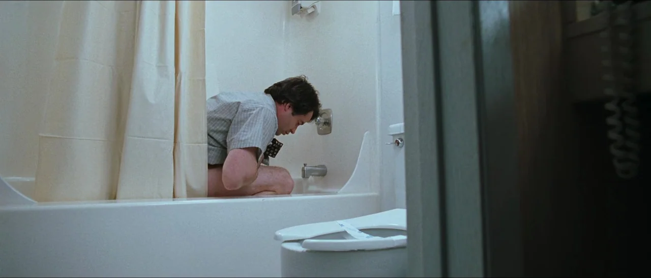 Election (1999) movie still: medium — A plain, cream-colored hotel bathroom features a tub, a shower curtain, and a toilet. A man in a patterned…