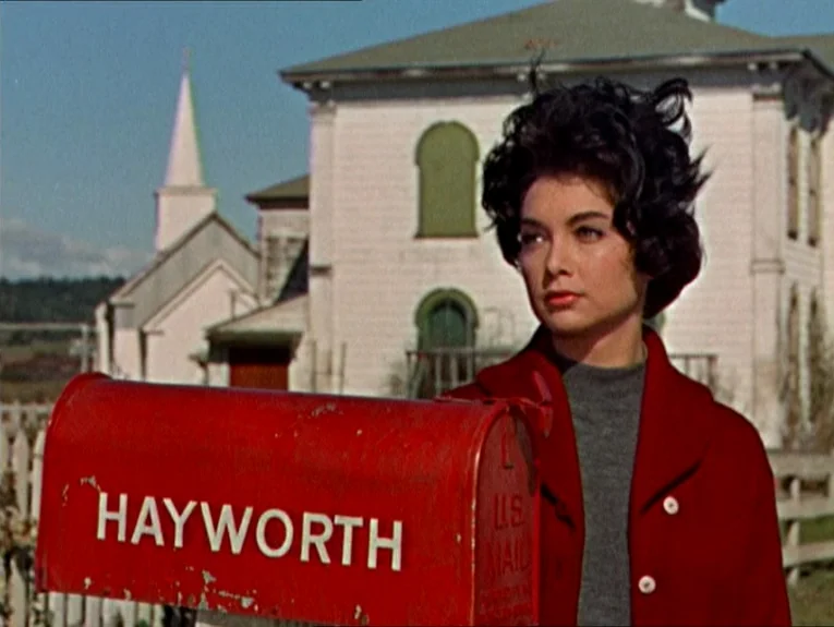 The Birds movie still: medium — A woman stands near a red mailbox labeled Hayworth in front of a building. The structure in the background features…