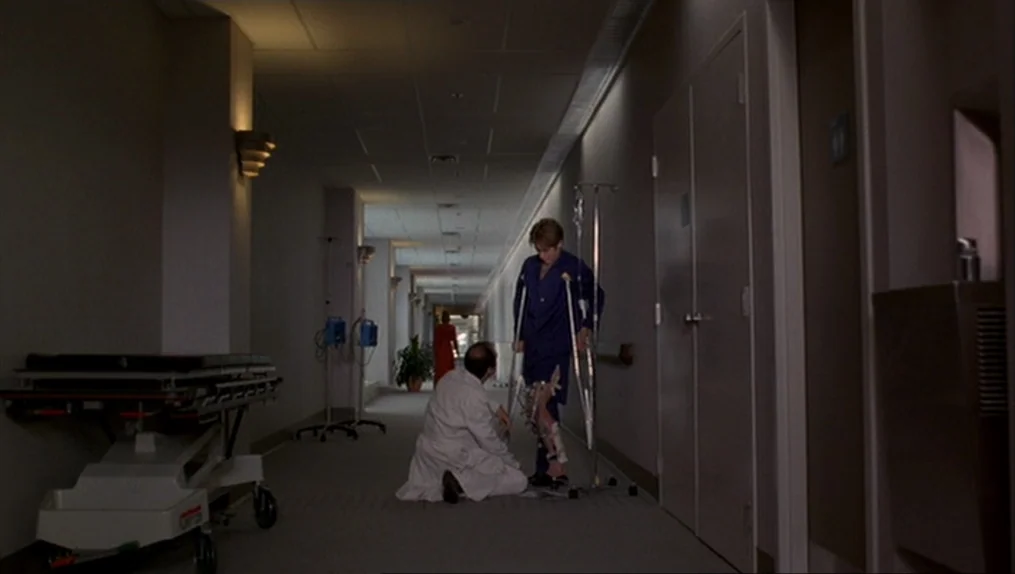 Crash (1996) movie still: wide — A long, dimly lit institutional hallway features linoleum floors and doors lining both walls. A doctor kneels to…