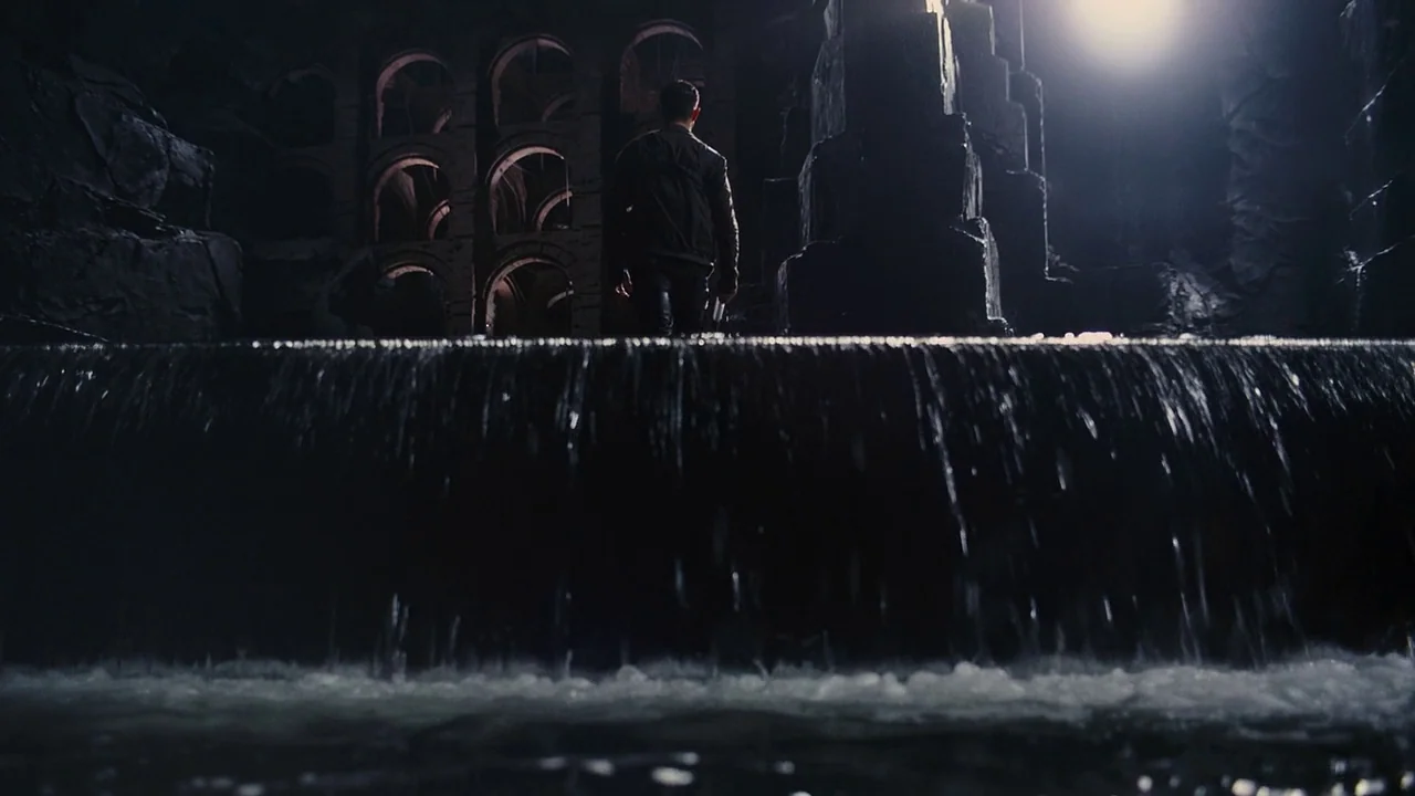 The Dark Knight Rises (2012) movie still: wide — A large dark stone sewer structure features arched walls and a flowing water ledge. A man stands on…