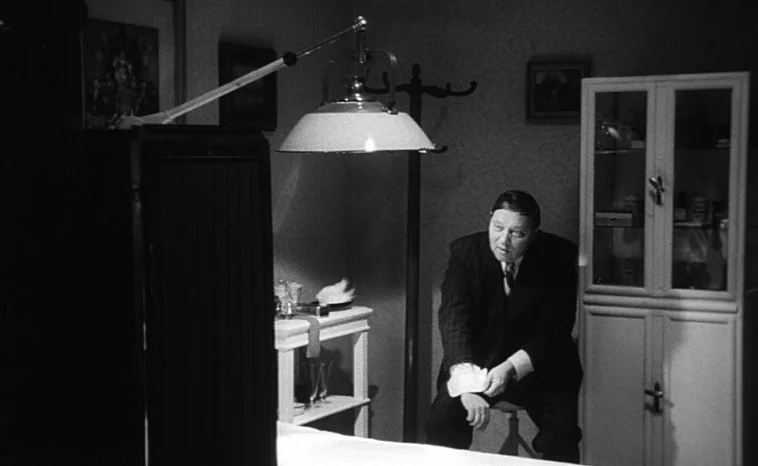 The Cremator (1969) movie still: medium — A sparsely furnished room contains a desk, a tall cabinet, and a coat rack. A man in a suit sits on a…