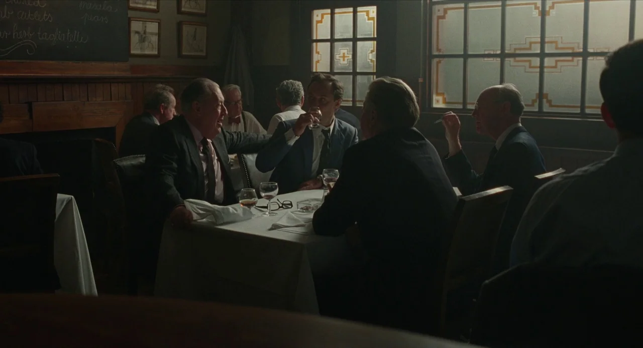 The Nest (2020) movie still: medium — A dimly lit, wood-paneled restaurant features men seated at tables with white tablecloths. A group of men in…