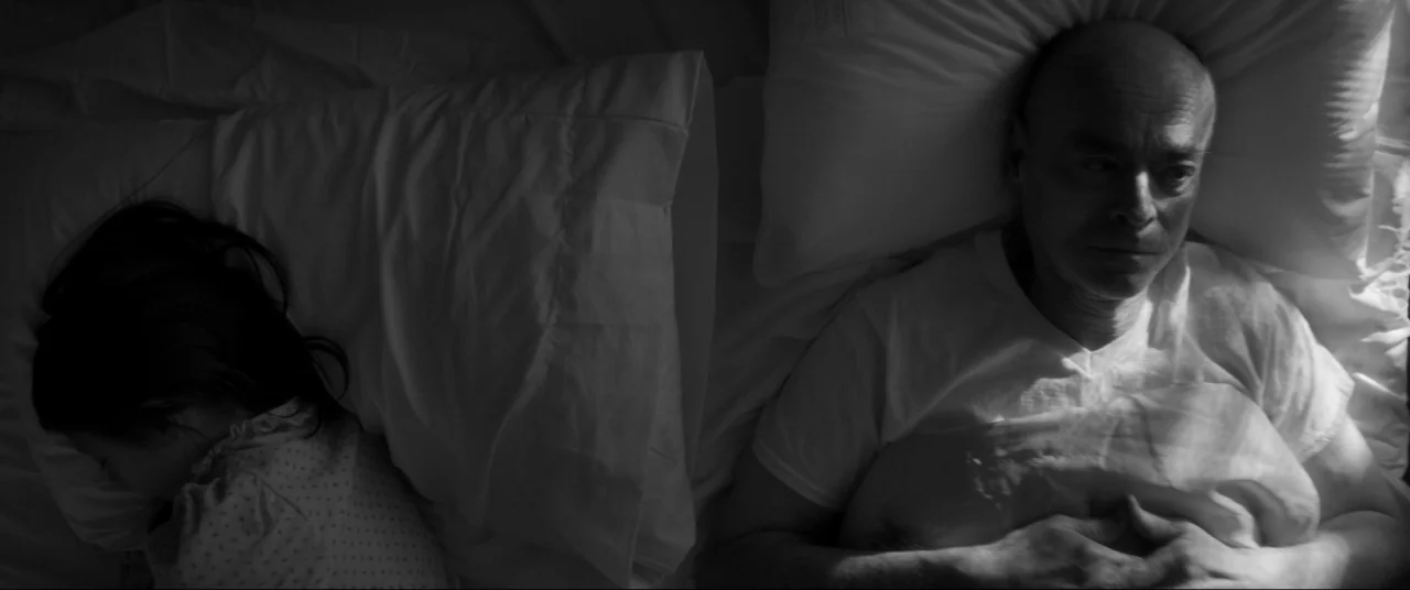 The Eyes of My Mother (2016) movie still: medium — Two people lie in a white bed under soft, high-contrast overhead light. An older man lies awake…
