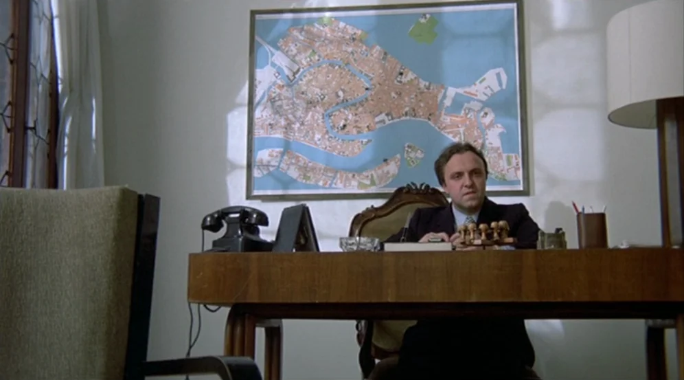 Don’t Look Now (1973) movie still: medium — A man sits behind a wooden desk in a room with white walls. A large detailed map of Venice hangs…