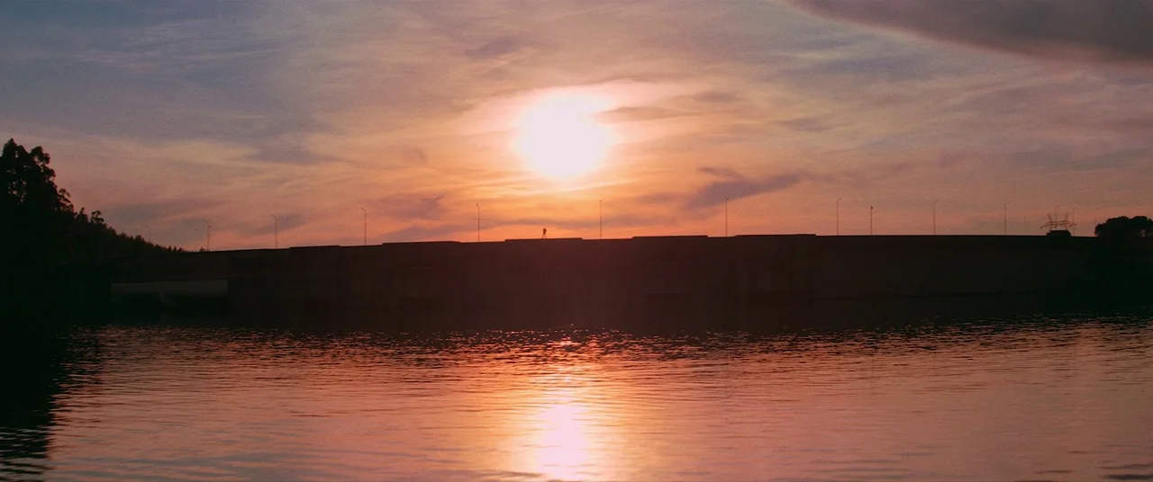 Colour Out of Space (2020) movie still: wide — A long, flat concrete dam structure spans across the frame against a sunset sky. The sun sets…