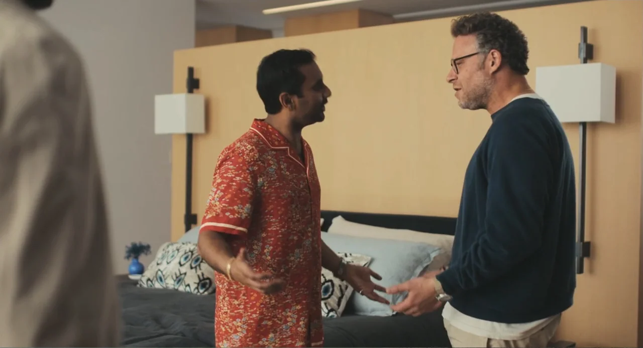 Good Fortune (2025) movie still: medium — A modern hotel room features a large light-colored headboard and patterned decorative pillows. Two men…