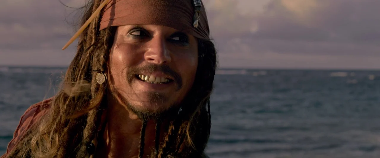 Pirates of the Caribbean: On Stranger Tides movie still: closeup — A man stands before a calm ocean during a sunset sky. The person grins while…