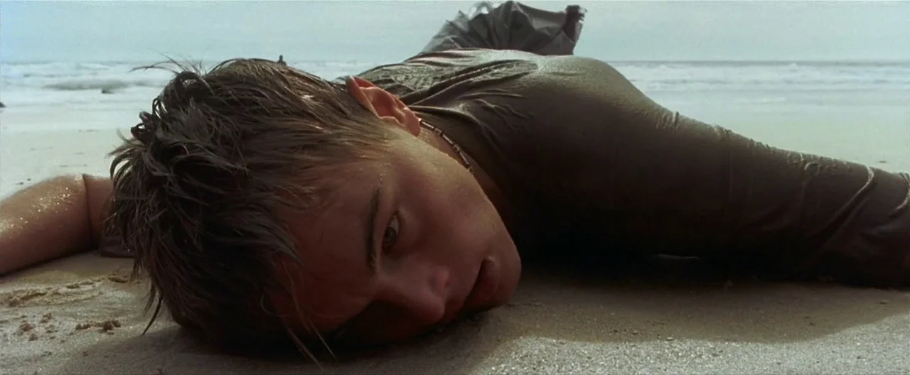 The Beach (2000) movie still: closeup — A wet sandy beach meets the ocean surf under a gray overcast sky. A young man lies prone in the sand with…