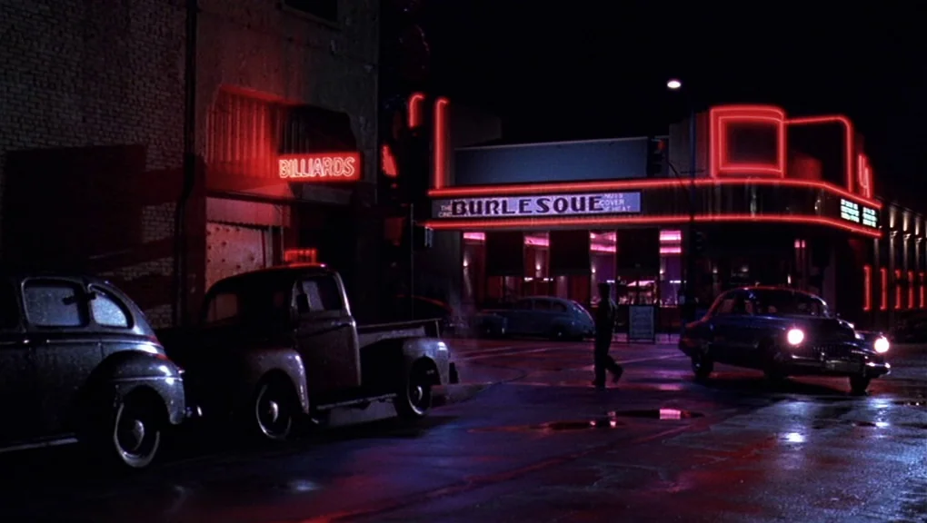 Bird (1988) movie still: wide — A dark, rain-slicked city street features a neon-lit burlesque theater and billiard hall. Vintage cars are parked…